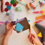 A person working on a scrapbook insert above a table with an assortment of crafting supplies on it, including paper and ribbon.