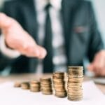 A businessperson gestures forward with their hand toward increasingly larger stacks of different coins.