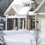 A tan, two-story home has a small front porch with a brown, wooden door. Snow covers the front yard and driveway.