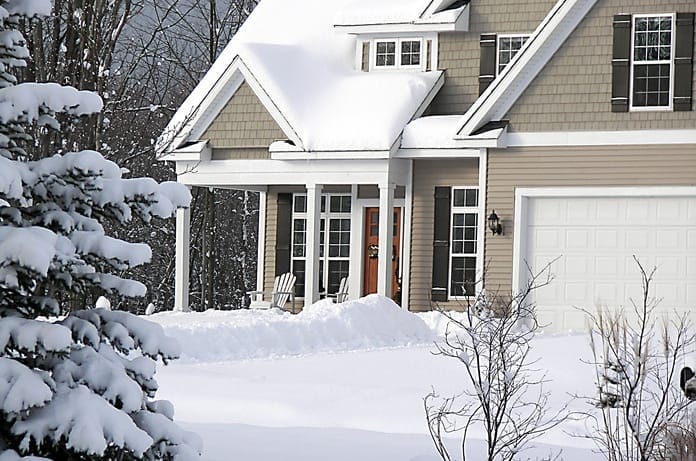 A tan, two-story home has a small front porch with a brown, wooden door. Snow covers the front yard and driveway.