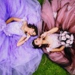 Two teens in elegant quinceañera gowns lie on grass, facing each other. One wears lavender, the other pink and black.