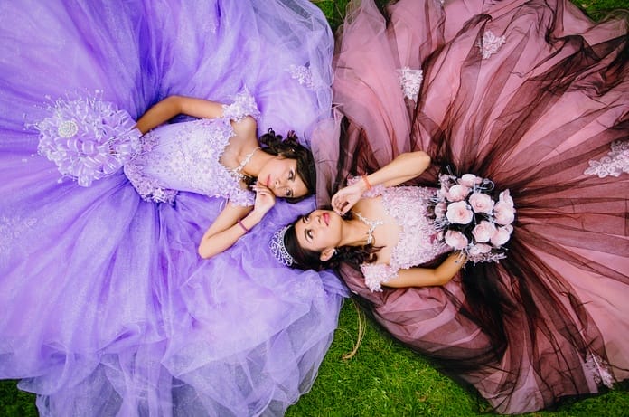 Two teens in elegant quinceañera gowns lie on grass, facing each other. One wears lavender, the other pink and black.