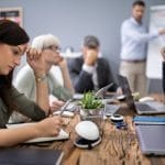 People sitting around a table with their hands on their heads and gaze downcast; in the background, a man points at a poster.