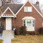 An old brick home with a white door, white windows, concrete steps, and a very inclined roof next to a tree.