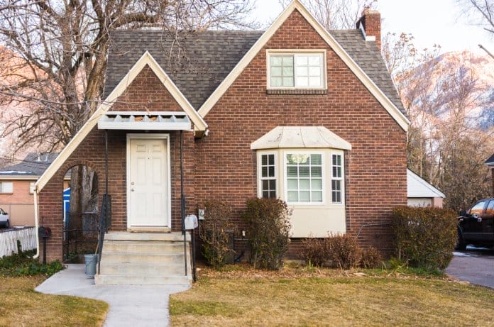 An old brick home with a white door, white windows, concrete steps, and a very inclined roof next to a tree.