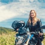 A woman smiles as she sits on her motorcycle while wearing a leather jacket. A large green hill sits behind her.
