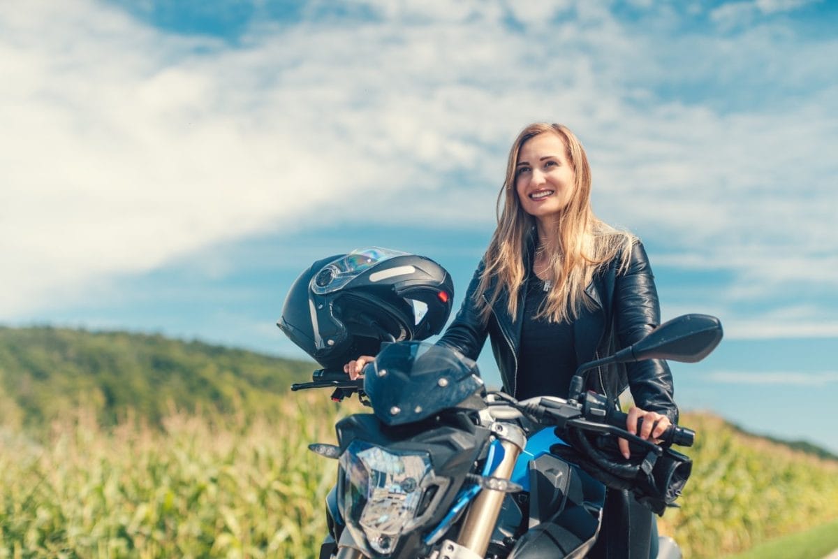 A woman smiles as she sits on her motorcycle while wearing a leather jacket. A large green hill sits behind her.
