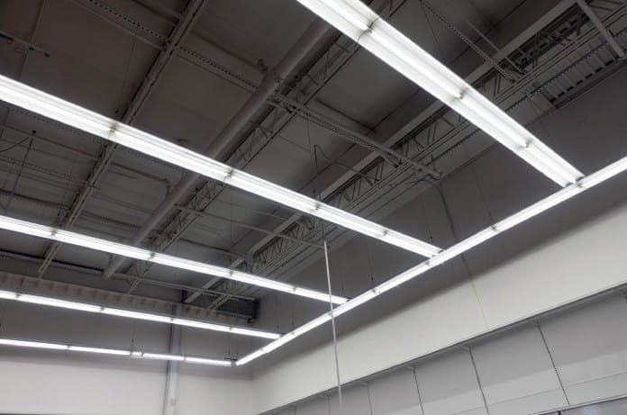 A close-up of white commercial strip lights hanging from an exposed ceiling of pipes and beams in a large retail store.