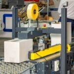 Home 15 A gray and yellow conveyor belt is pushing a white cardboard box through a gray automatic taping machine.